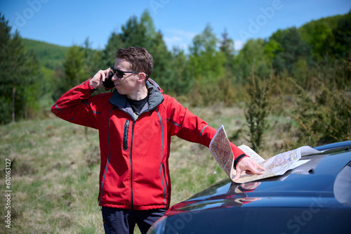 A man leans against a car, holding a map and cellphone in his hands, looks for directions. The traveler behind the wheel has become disoriented and is using the map to determine their next route.