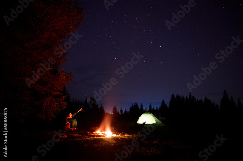 A young female traveler sits by the campfire near her tent  enjoying a moment of relaxation beneath the beautifully starry sky. As the crackling flames provide warmth and comfort