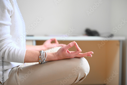 Businesswoman meditating in the office while sitting on the chair. Side crop view. Lotus pose. Practicing meditation on office chair with hands in mudra gesture. Stress relief during freelance work.