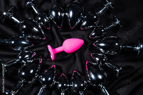 Pink silicone butt plug among many metal ones on a black silk sheet. 
