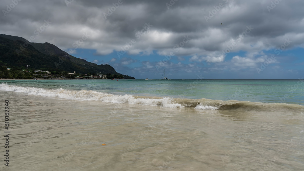 A wave of turquoise ocean rolls towards the shore, twisting and mixing with the sand of the beach. A green hill against a background of blue sky and clouds. Seychelles. Mahe. Beau Vallon.