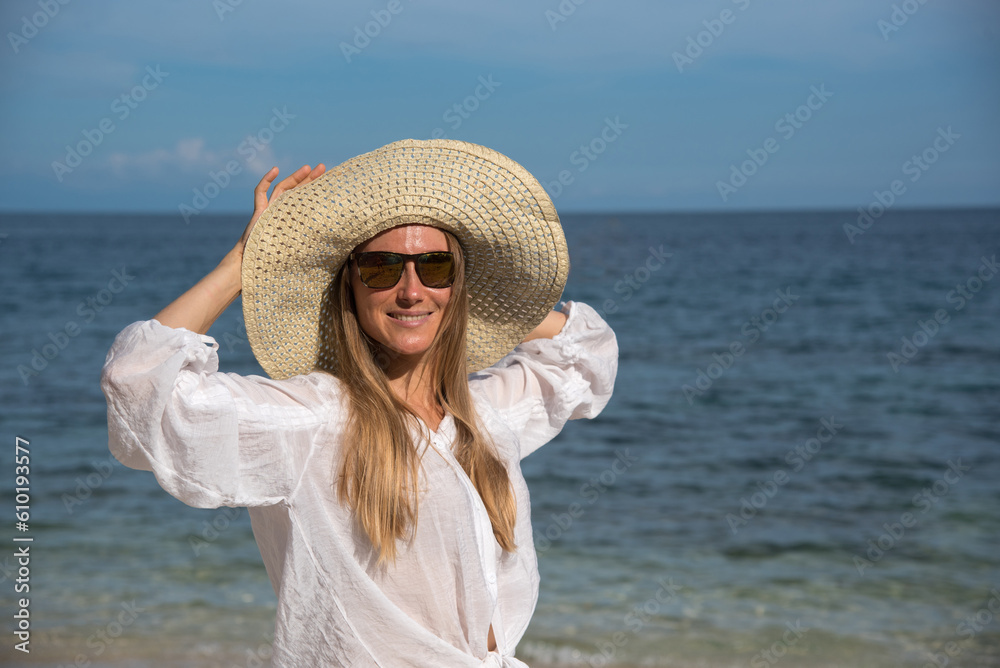 A happy blond woman with sunglasses is standing by the sea. She is touching her straw hat. A girl with long hair and a white shirt spends her vacation on the beach. Holidays to other countries