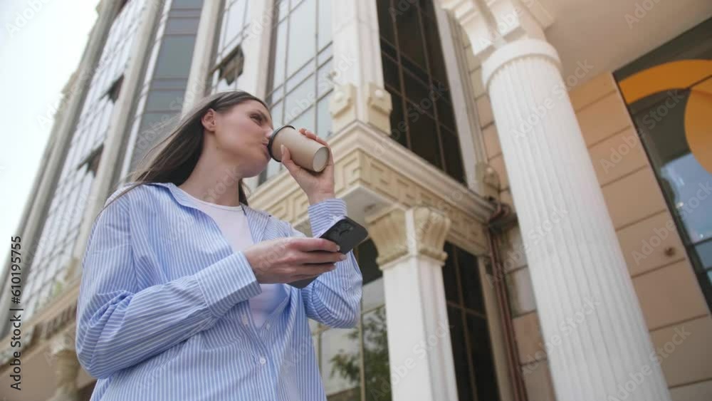 Beautiful young woman drinking takeaway coffee while using mobile phone near building, low angle view. Slow motion effect Beautiful young woman drinking takeaway coffee while using mobile phone near building, low angle view. Slow motion effect