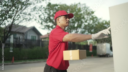 A parcel delivery worker dressed in a red uniform is holding a parcel box and ringing the bell for the recipient. contact the receiver in front of the house.