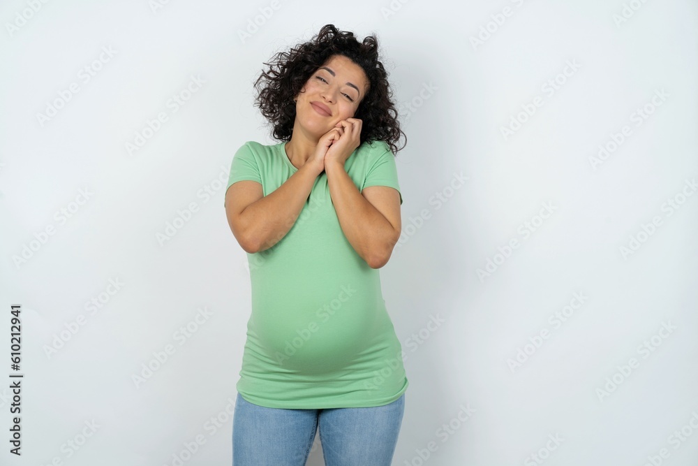 Charming serious young pregnant woman wearing green t-shirt over white background keeps hands near face smiles tenderly at camera