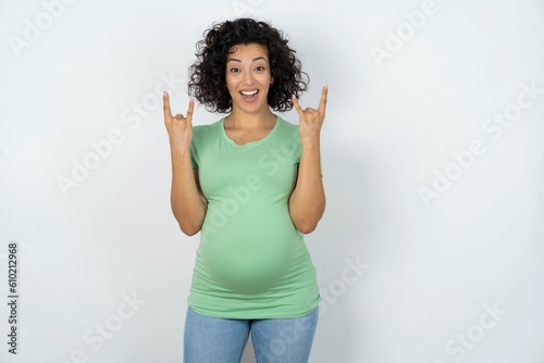 young pregnant woman wearing green t-shirt over white background makes rock n roll sign looks self confident and cheerful enjoys cool music at party. Body language concept.