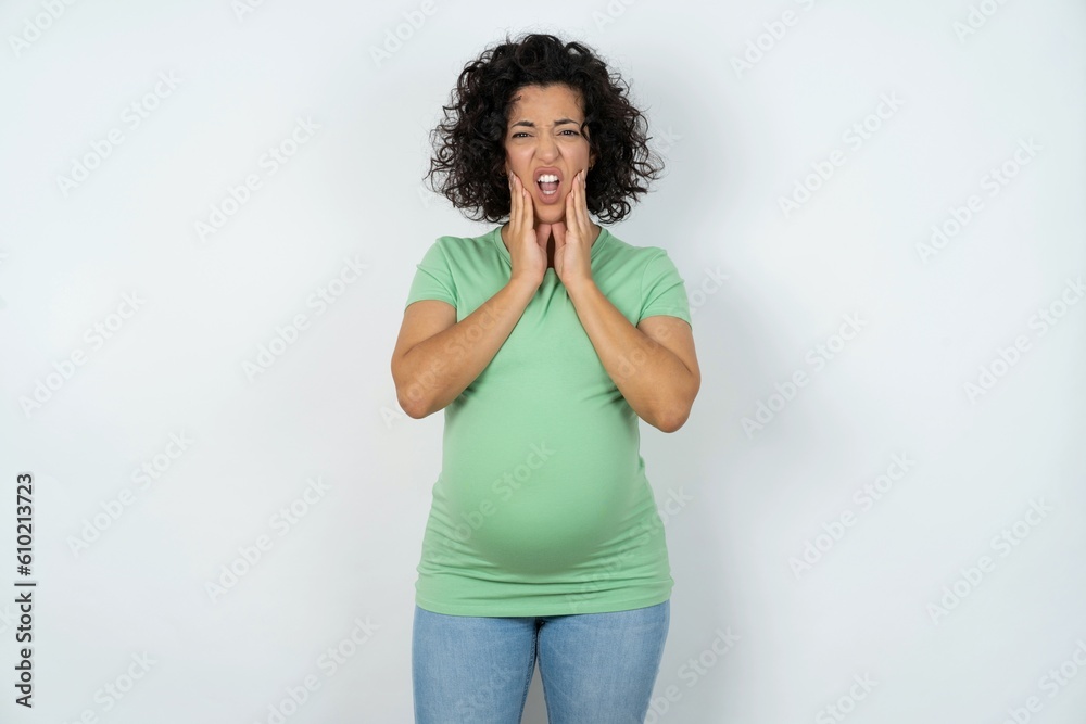 Upset young pregnant woman wearing green t-shirt over white background touching face with two hands Upset young pregnant woman wearing green t-shirt over white background touching face with two hands