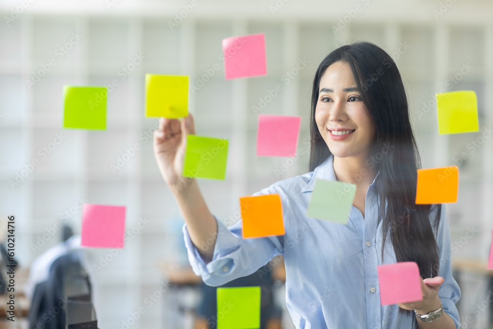 Smiling business Asian people thinking and use sticky notes on glass wall in office with her colleague in background.
Smiling business Asian people thinking and use sticky notes on glass wall in office with her colleague in background.