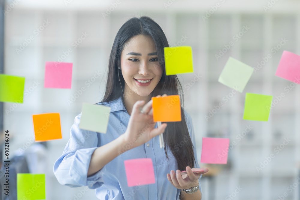 Smiling business Asian people thinking and use sticky notes on glass wall in office with her colleague in background.
Smiling business Asian people thinking and use sticky notes on glass wall in office with her colleague in background.