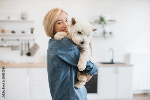 Obraz na plátně Portrait of smiling blonde lady with West Highland White Terrier in arms posing on background of spacious kitchen