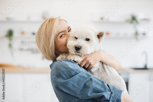 Obraz na plátně Portrait of smiling blonde lady with West Highland White Terrier in arms posing on background of spacious kitchen