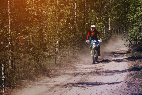 motorcycle racer on a sports off-road enduro motorcycle rides fast on a dusty road through the forest in a race on a sunny day