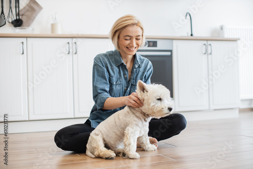 Happy caucasian female adult in casual clothes rubbing ears of small terrier while sitting cross-legged in kitchen. Smiling slim woman making her adorable furry friend feel relaxed during day indoors.