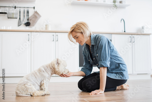 Beautiful blonde lady in cozy clothes feeding small furry pet from hand while staying at home on weekend. Attentive female keeper giving animal favourite treat for successfully followed command. photo