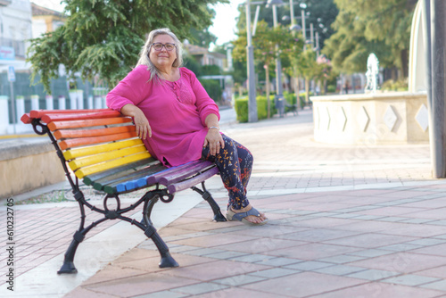 older woman with white hair and pink shirt sitting on a bench painted with the colors of the LGTBIQ flag looking at the camera. © JOSEANTONA