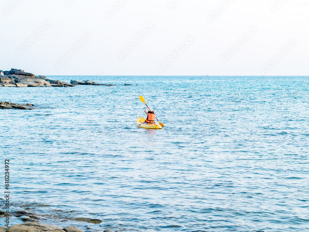 Back of Asian woman in life jacket and cap kayaking on yellow kayak boat with using paddle on the sea. Happy female having fun activity on seascape view background, Holiday trip vacation.