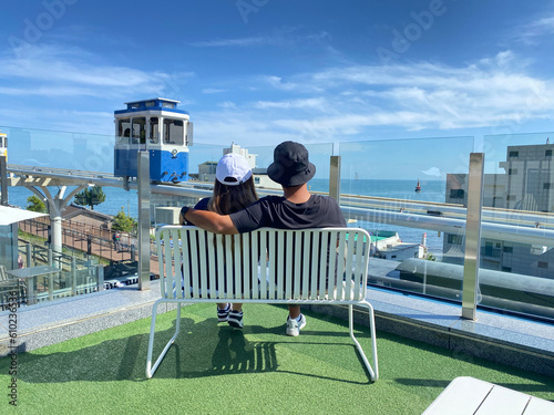 Couple travellers sitting and looking at the Sky Capsule Tram Haeundae Blue Line at Mipo Station for traveling at Haeundae Beach Park. photo