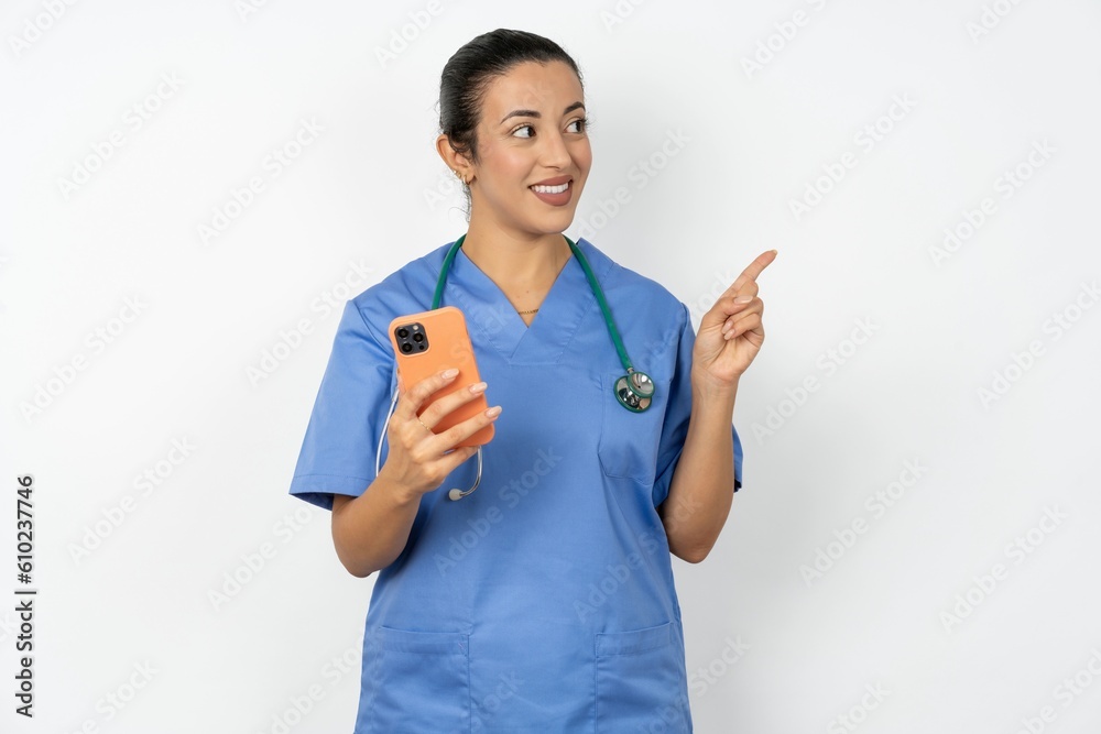 Wow!! excited Young doctor woman wearing blue uniform over isolated background showing mobile phone with open hand gesture Wow!! excited Young doctor woman wearing blue uniform over isolated background showing mobile phone with open hand gesture