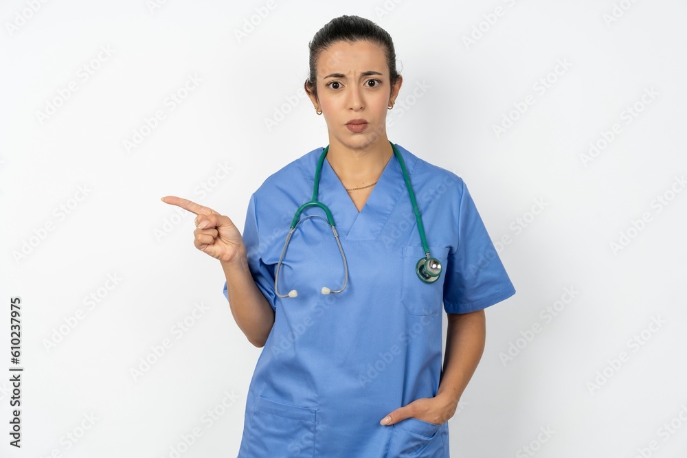 Serious Young doctor woman wearing blue uniform over isolated background smirks face points away on copy space shows something unpleasant. Look at this advertisement. Big price concept Serious Young doctor woman wearing blue uniform over isolated background smirks face points away on copy space shows something unpleasant. Look at this advertisement. Big price concept