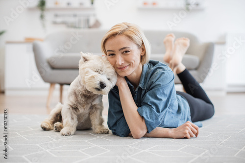Leinwand Poster Full length portrait of lovely smiling woman in denim shirt caressing adorable little Westie on kitchen floor of apartment