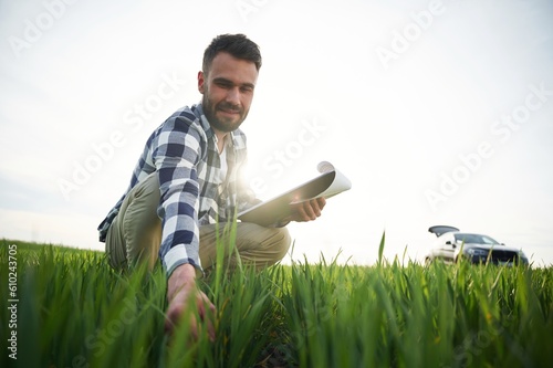 Car is behind. With notepad and wheat in hands. Handsome young man is on agricultural field