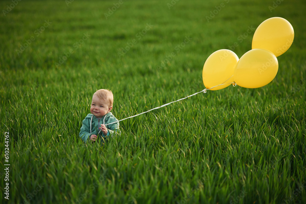 With yellow balloons in hands. Cute little baby boy is in the agricultural field at daytime