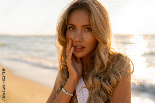  Close up portrait of beautiful blond woman in sexy swimwear posing on the beach in sunset light. Prtfect wavy hairs, tan skim body. Summer tropical mood.