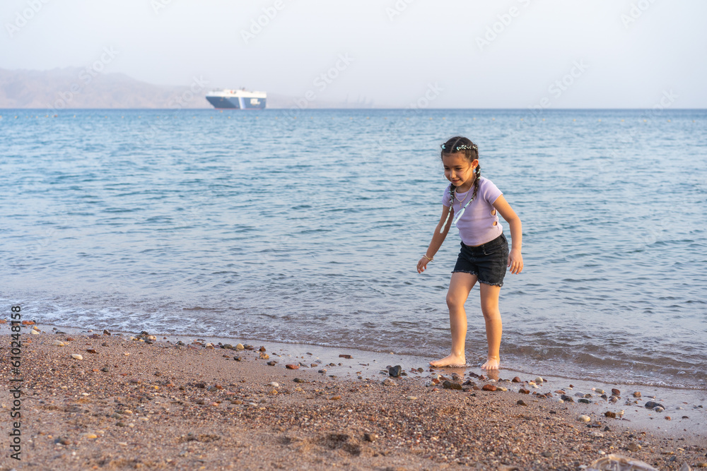 Cute little child playing on the beach. Little beautiful girl with braided hair walking barefoot on the seashore.