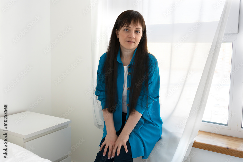 Young happy woman wearing blue shirt sitting on the windowsill in a new apartment. Mortgage concept. Young happy woman wearing blue shirt sitting on the windowsill in a new apartment. Mortgage concept.