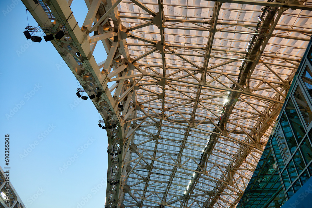 Bottom view of the stadium roof with cloudless blue sky. Floodlights are placed high on the metal structure of the roof of a large stadium.