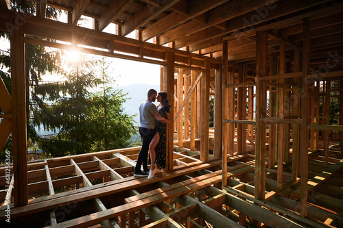 Man and woman examining their future wooden frame dwelling nestled in the mountains near forest. Youthful couple at construction site in early morning. Concept of contemporary ecological construction.