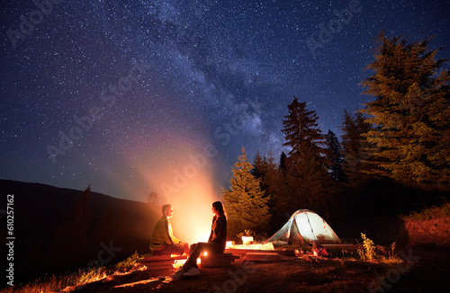 Overnight camping escapade in mountains, under glistening stars. Man and woman, avid explorers, rejuvenate by crackling fire and tent, under the mesmerizing night sky adorned with the Chumack Trail.