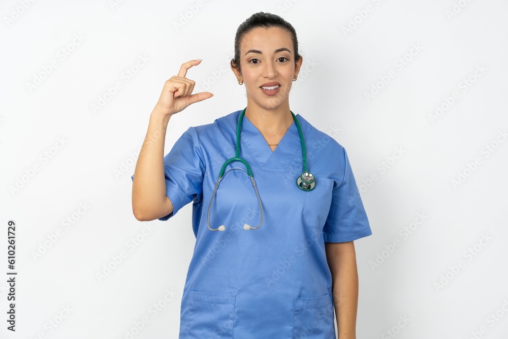 Beautiful doctor woman standing over white studio background smiling and confident gesturing with hand doing small size sign with fingers looking and the camera. Measure concept Beautiful doctor woman standing over white studio background smiling and confident gesturing with hand doing small size sign with fingers looking and the camera. Measure concept