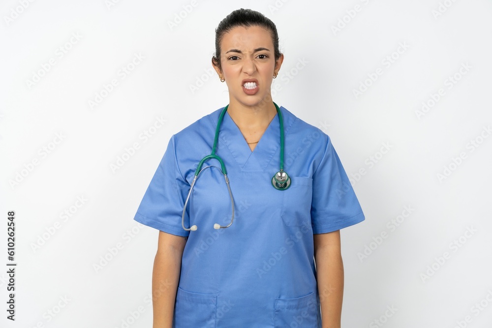 Beautiful doctor woman standing over white studio background keeps teeth clenched, frowns face in dissatisfaction, irritated because of much duties. Beautiful doctor woman standing over white studio background keeps teeth clenched, frowns face in dissatisfaction, irritated because of much duties.