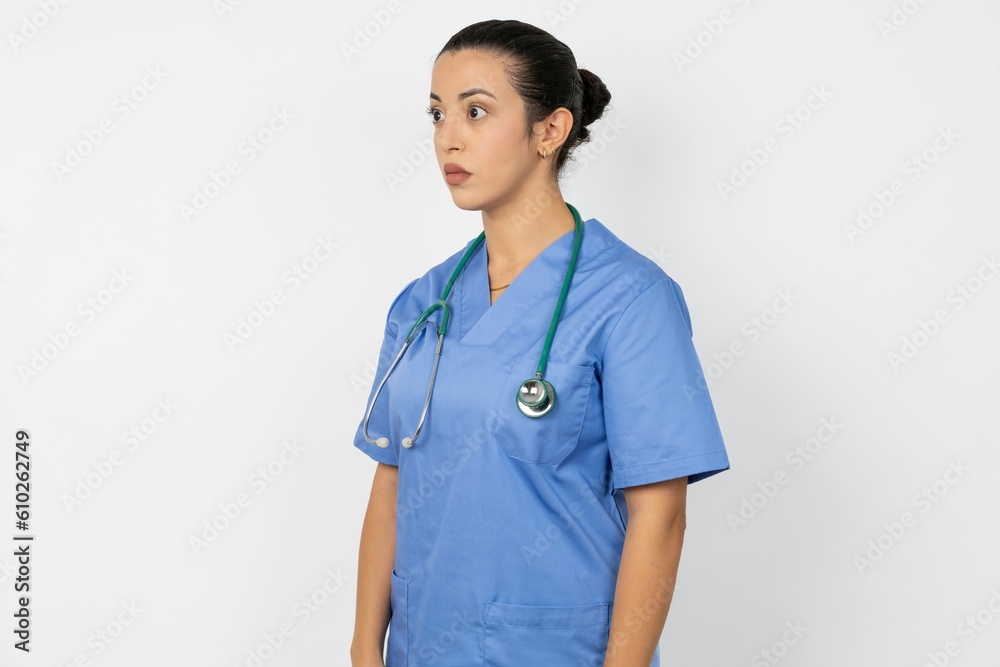 Beautiful doctor woman standing over white studio background stares aside with wondered expression has speechless expression. Embarrassed model looks in surprise