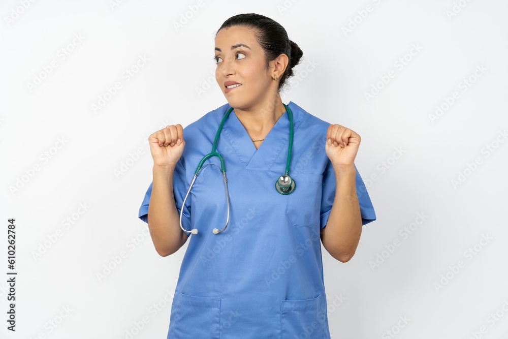 Beautiful doctor woman standing over white studio background clenches fists and awaits for something nice happened looks away bites lips and waits announcement of results Beautiful doctor woman standing over white studio background clenches fists and awaits for something nice happened looks away bites lips and waits announcement of results