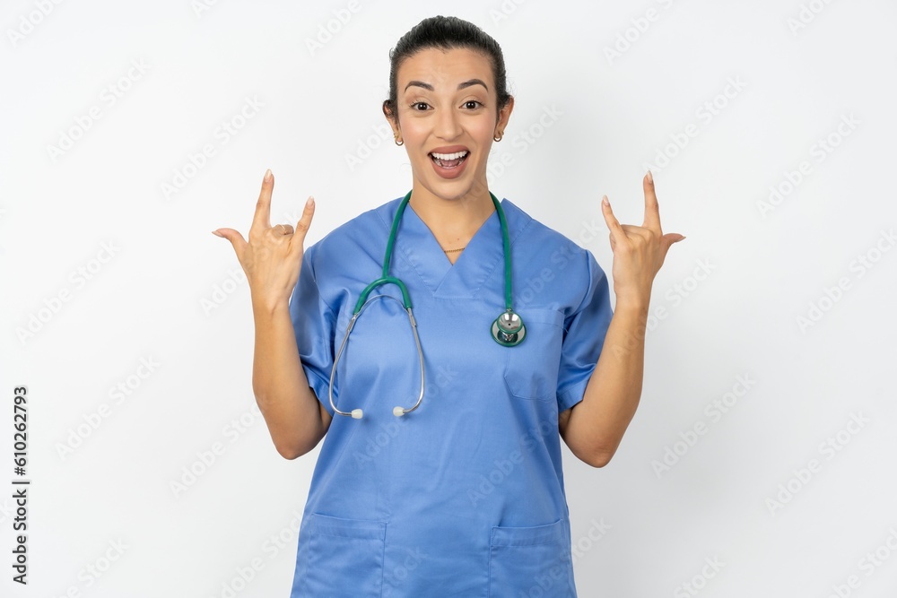 Beautiful doctor woman standing over white studio background makes rock n roll sign looks self confident and cheerful enjoys cool music at party. Body language concept. Beautiful doctor woman standing over white studio background makes rock n roll sign looks self confident and cheerful enjoys cool music at party. Body language concept.