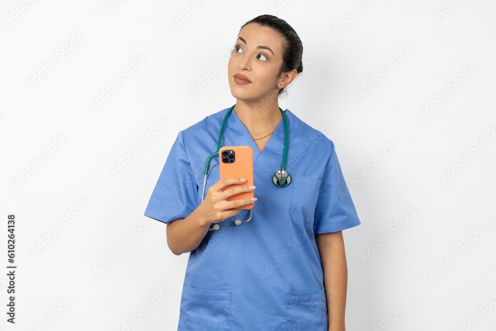 Beautiful doctor woman standing over white studio background holds mobile phone uses high speed internet and social networks has online communication. Modern technologies concept