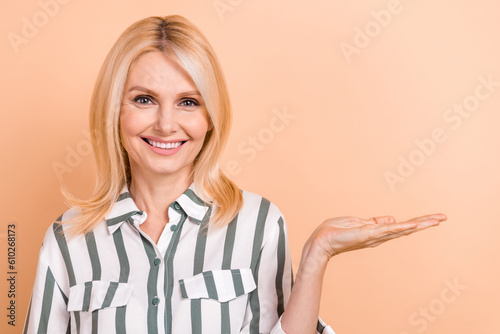 Portrait of positive senior woman with blond hairdo wear striped formalwear palm demonstrate object isolated on beige color background