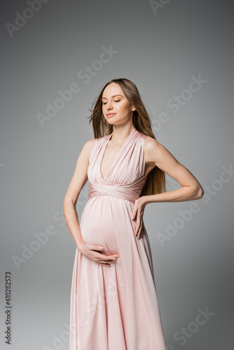 Fashionable long haired and pregnant woman with closed eyes touching belly while posing in pink dress isolated on grey, elegant and stylish pregnancy attire, sensuality, mother-to-be