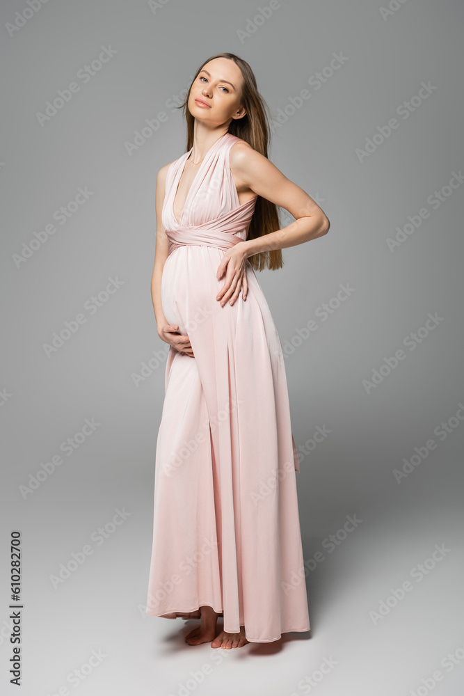 Trendy and fair haired pregnant woman in pink dress looking at camera while posing on grey background, elegant and stylish pregnancy attire, sensuality, mother-to-be