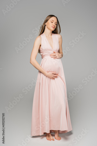 Full length of barefoot and fair haired pregnant woman in elegant pink dress touching belly and standing with closed eyes on grey background, elegant and stylish pregnancy attire