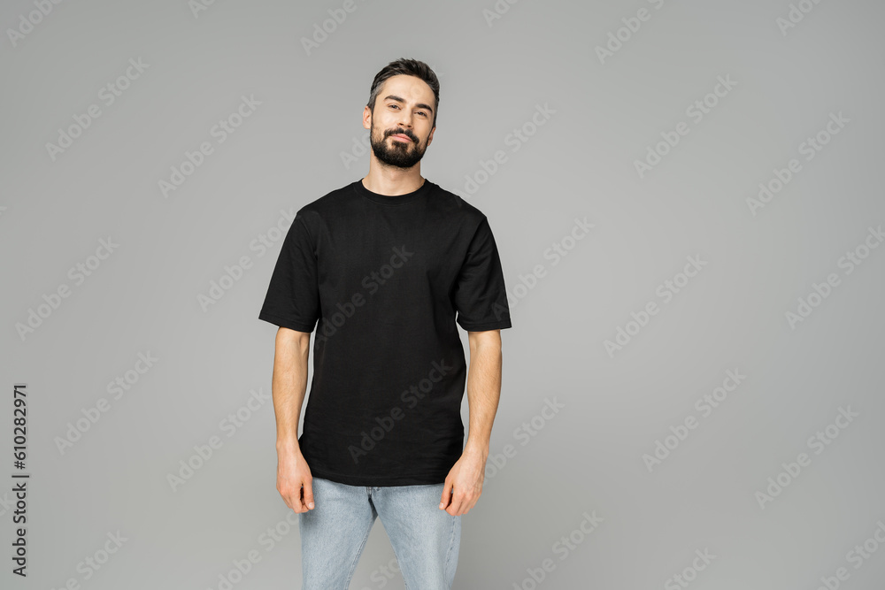 Confident and relaxed bearded and brunette man in black t-shirt and jeans looking at camera while standing isolated on grey, masculine beauty concept, fashion-forward Confident and relaxed bearded and brunette man in black t-shirt and jeans looking at camera while standing isolated on grey, masculine beauty concept, fashion-forward