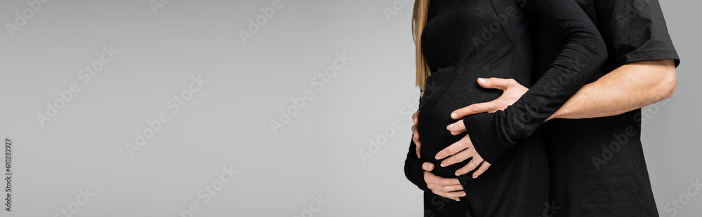 Cropped view of man in t-shirt hugging and touching belly of stylish pregnant wife in black dress and standing together isolated on grey, growing new life concept, banner