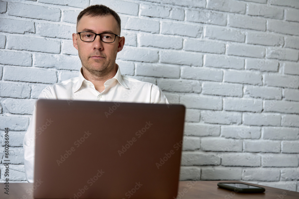 Remote work of a freelancer man in glasses using a laptop and smartphone. Man with glasses and a white shirt looks at the laptop screen sitting in the office Remote work of a freelancer man in glasses using a laptop and smartphone. Man with glasses and a white shirt looks at the laptop screen sitting in the office