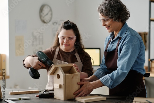 Fototapeta Girl with down syndrome using drill to make birdhouse with woman helping her wit