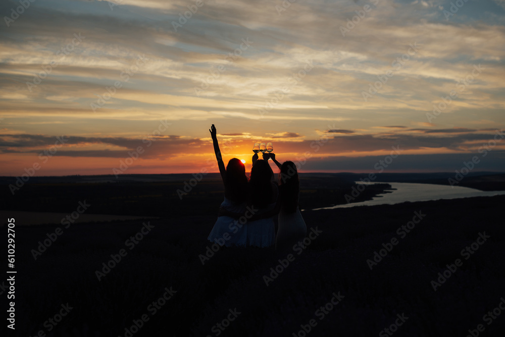 Lovely ladies drinking wine in lavender violet field at sunset. Summer happy mood. Girlfriends relaxing on summer sunset with river on the background.