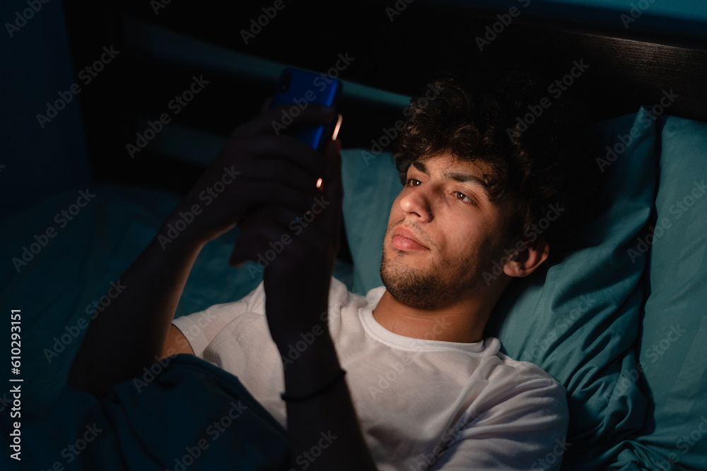 Insomnia, addiction concept. Sleepy exhausted man lying in bed using smartphone, can not sleep. Handsome guy in bed scrolling through social networks on mobile phone at night in dark - obrazy, fototapety, plakaty 