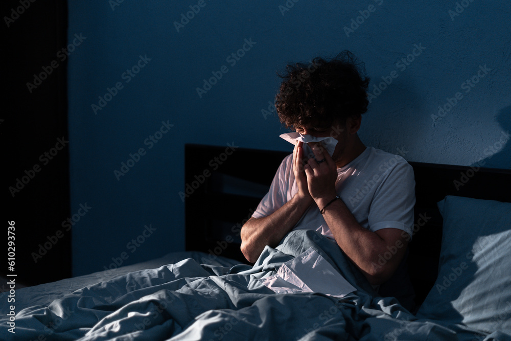 Handsome young man suffering cold and flu trying to sleep at night, guy sneezing into a tissue in bedroom. Illness colds or flu. Allergy or health issue concept. - obrazy, fototapety, plakaty 