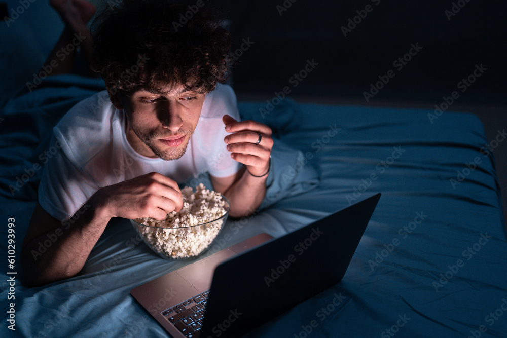 Young man eating popcorn and watching movie on laptop computer at night at home while lying on bed - obrazy, fototapety, plakaty 